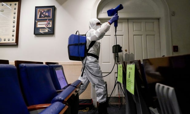 A person wearing a white boiler suit and mask carries a blue backpack and holds a spray attachment as they walk past rows of blue upholstered flip chairs