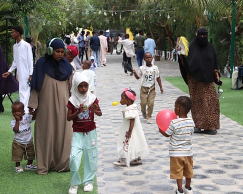 Children with balloons and adults walk on a concrete path surrounded by grass and trees