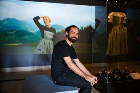 A bearded man sits in front of a photo from the film Sound of Music next to a mannequin in a dirndl