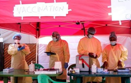People in medical gowns and masks stand in a tent under a handwritten sign saying ‘vaccination’.