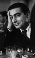 Black and white photograph of Robert Capa, head resting agains his hand, smiling slightly with a wine glass in front of him
