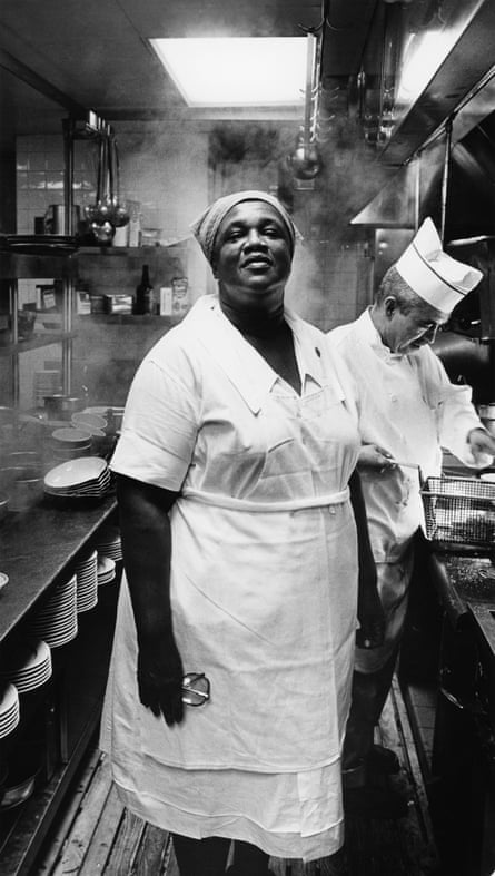 a woman in white uniform stands in kitchen looking ahead as a man cooks next to her