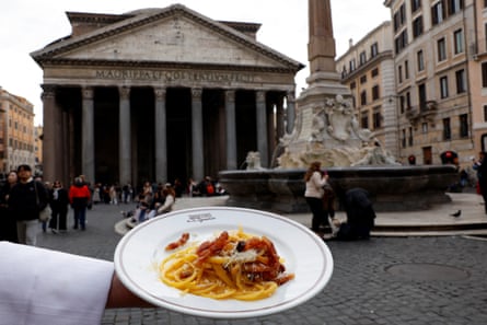 A the arm of a white-coasted waiter holds a of pasta with the colonnaded building in the background