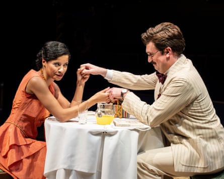 Boadicea Ricketts and James Sheldon sit across a table from each other, holding hands. Ricketts wears an orange dress and Sheldon wears a cream striped suit and glasses