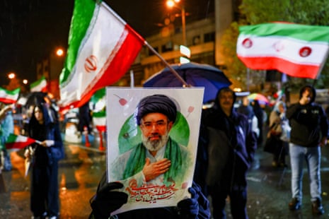 A woman covers her face with a poster of Mojtaba Khamenei while waving an Iranian flag.