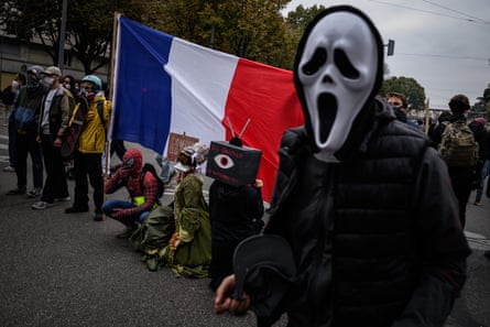 At a Lyon protest, a person in a ghost mask stands in a crowd near a large French flag