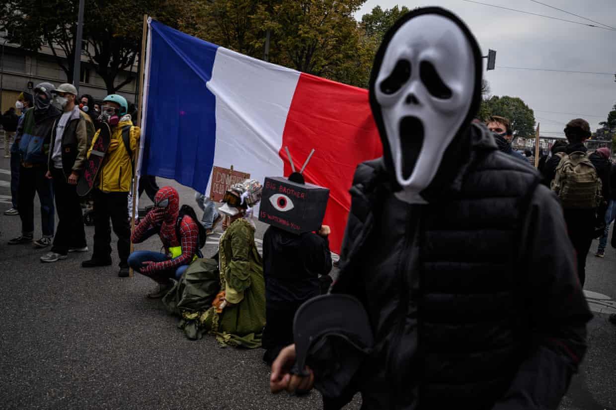 At a Lyon protest, a person in a ghost mask stands in a crowd near a large French flag