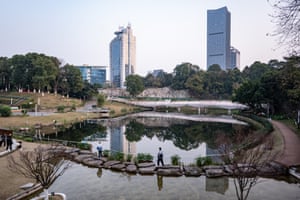 People walk along stepping stones across a lake in a park with two skyscrapers in the background