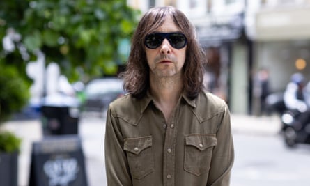 Bobby Gillespie of Primal Scream, with sunglasses and long hair, wearing a cowboy-style long-sleeved T-shirt, looking serious in the street