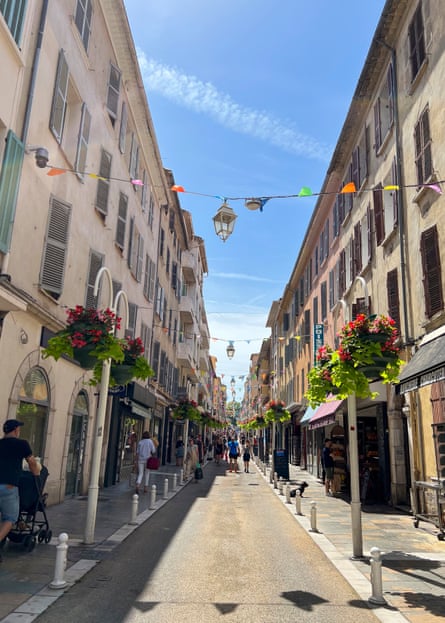 A narrow street with high, shuttered buildings and flowers hanging from lamp-posts