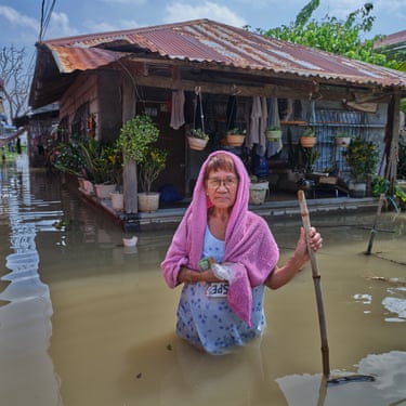 A woman stands in a flood street up to her waist holding a stick and with a pink towel over her head and a wooden house with a tin roof behind her