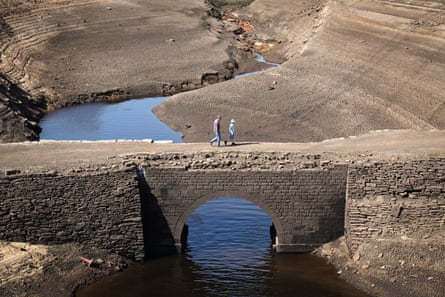 People walk on a bridge over a reservoir with low water level.