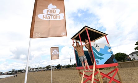 Two women in a makeshift hut on the beach, their hands to their foreheads looking out, with signs nearby saying ‘poo watch’