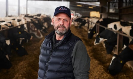 Michael Oakes at his farm in Worcestershire. He is standing in an indoor shed with rows of cattle on either side.