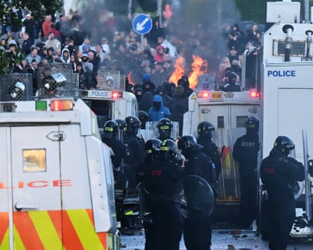 Police stand guard next to vehicles, with rioters and fires in the distance