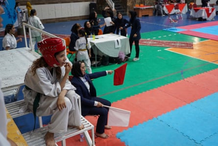 A woman in a headscarf sits holding two flags at the edge of a sports mat, while a younger woman in a karate gi and helmet perches beside her.