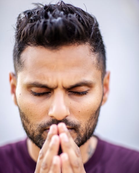 A young Indian man with light black beard and gelled black hair and a dark purple T-shirt closes his eyes and seems to scowl intently as he raises his folded hands to his lips.