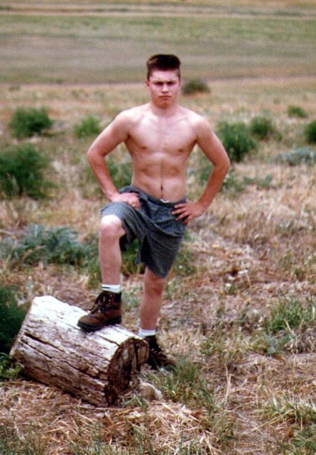 Jason Whiter, at 18 with a short cropped haircut, stands shirtless outside, looking tough, with a knee up on a log.