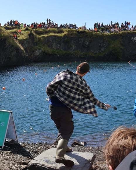 A man throws a stone into water at the World Stone Skimming Championships, crowd watching from cliffs above