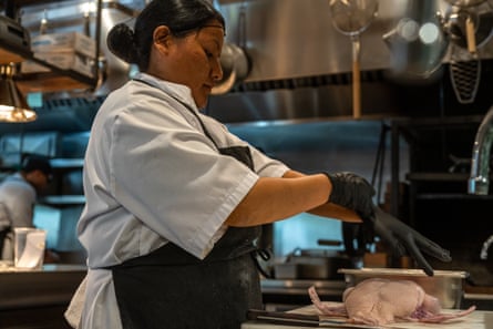 A woman wearing an apron and standing in a professional kitchen pulls on black food service gloves. In front of her, on a white cutting board, sits a raw duck carcass.