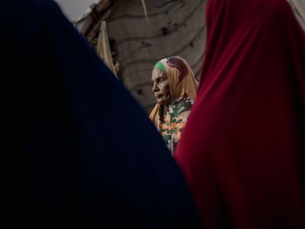 A displacement camp resident, stands outside her shelter, framed by figures in dark and red fabric