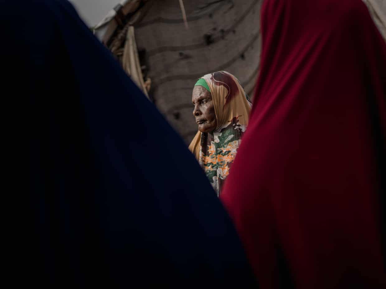 A displacement camp resident, stands outside her shelter, framed by figures in dark and red fabric