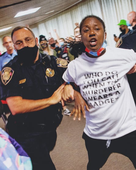 a police officer wearing a face mask detains a shouting person inside a building who wears a shirt that says “who do you call when the murderer wears a badge?”