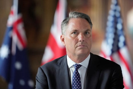 A man with grey hair sits before three flags.