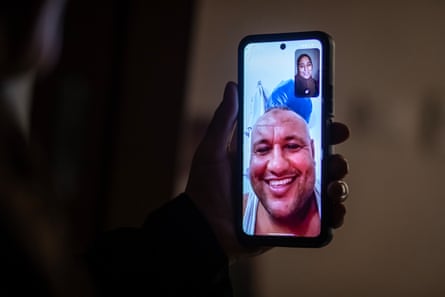 Wijdan, standing in the dark, holds up her phone screen to show an illuminated image of Hatem as they make a video call. He is smiling on the screen.