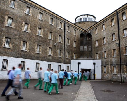 Prisoners wearing blue tops and green trousers walk from a yard into a Victorian building.