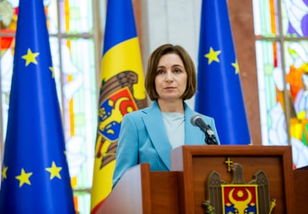 Maia Sandu speaks in the presidential palace in Chisinau, Moldova: she stands in front of flags including the EU flag. She is in her early 50s and has straight brown hair in a chin-length bob; she wears a pale blue jacket.
