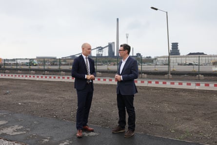 Two men in black suits standing on industrial land near steelworks visible in distance.