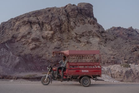 A man rides a red three-wheeled motorbike truck past a large rocky mountain on the island