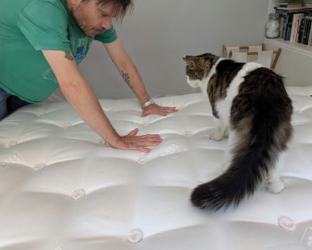 A man in a green T-shirt presses his hands into the Millbrook Wool Luxury 4000 mattress as a hat stands watching him