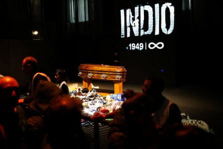 People stand in shadow in front of a small wooden coffin, which is illuminated with lettering behind it that reads Indio: 1949, plus an infinity symbol. Flowers and tributes are heaped in front of the coffin.