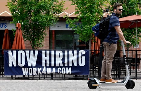 A man rides an electric scooter past a 'Now hiring' sign outside a business.
