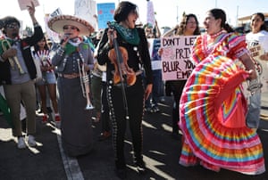 Two women, one in a dress and one holding a violin, smile at each other as a woman in a sombrero holding a saxophone talks into a microphone
