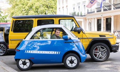 Jasper Jolly looks out of window of the blue Microlino electric car which is parked next to a larger vehicle yellow vehicle