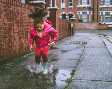 A little girl in a pink raincoat and green boots jumps in a puddle, hair flying up