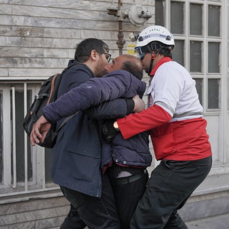 Two men stand either side of the collapsed man. The man on the right has his arms around him as they bring him to his feet
