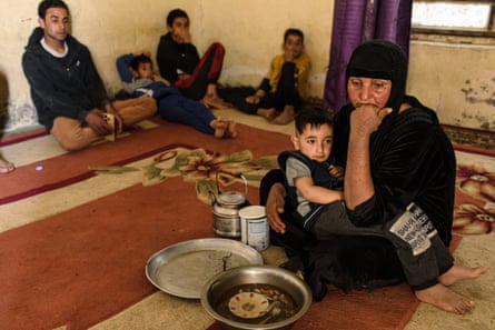 A woman sits cross-legged on a carpet with a baby boy on her lap, in front of dishes of food. Behind her, four boys sit against the wall