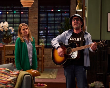 Lee plays guitar in an Oasis T-shirt while singing; Lucy, in a green cardigan, looks on, unimpressed.