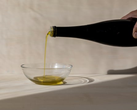 A hand can be partially seen holding a black bottle of olive oil and pouring into a small glass bowl. The photo is taken from the side and clearly shows the liquid in focus. Natural daylight has been used and is against a neutral background.