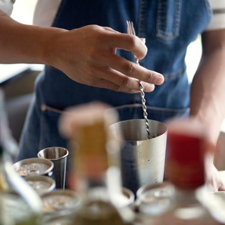 Male in denim apron using swizzle stick in cocktail shaker surrounded by cocktail making paraphernalia