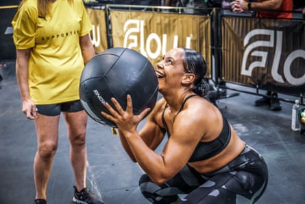 A woman squats down holding a medicine ball while someone wearing a yellow T-shirt looks on, perhaps checking that the first woman’s squat is low enough