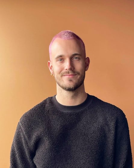 A man with cropped purple-dyed hair, wearing black jumper, standing in front of an orange background.