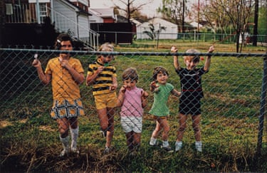 Five children of different ages standing behind a wire fence with a huge garden and houses behind them