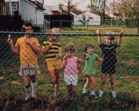 Five children of different ages standing behind a wire fence with a huge garden and houses behind them