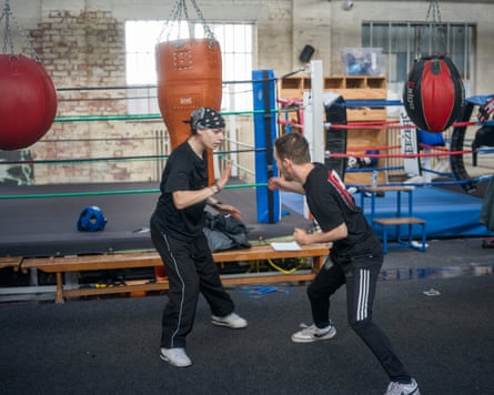 A young woman and a young man sparring in a boxing gym next to a ring and punchbags