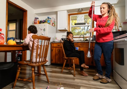 A woman sweeps in a kitchen and her two daughters sit in chairs eating lunch